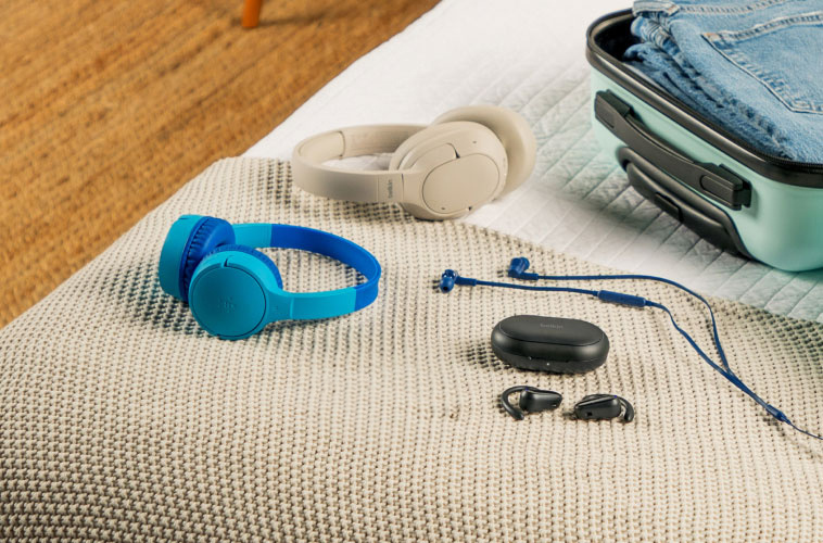 A variety of Belkin headphones and earbuds&mdash;including over-ear, on-ear, and wired models&mdash;neatly arranged on a bed next to an open mint green suitcase packed with clothes.